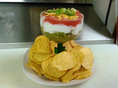 A layered dip in a glass cup topped with green onions and cheese, surrounded by a pile of tortilla chips.