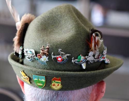 A close-up of a green hat adorned with various pins and badges, featuring feathers and colorful emblems.