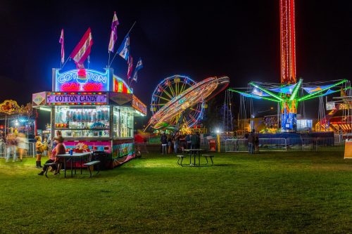 A vibrant carnival scene at night featuring a cotton candy stand, colorful rides, and illuminated attractions.
