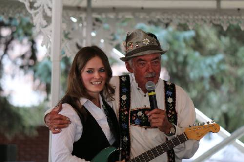 A man in traditional attire holds a microphone while embracing a woman with a guitar, both smiling on stage.