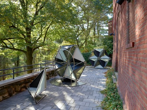 Three geometric sculptures in a park-like setting, surrounded by trees and a brick building.