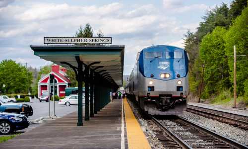 A train approaches the White Sulphur Springs station, surrounded by greenery and a quaint building in the background.