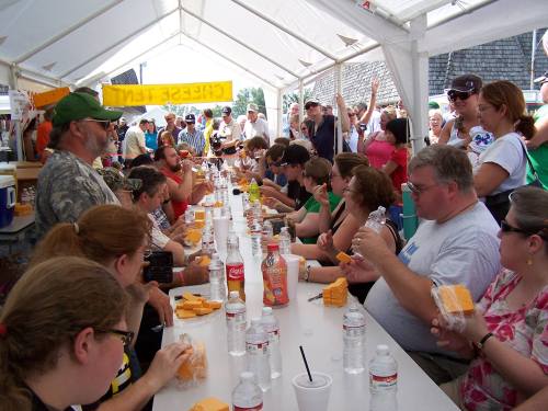 A crowd gathers under a tent, watching participants eat cheese at a contest, with drinks and snacks on the table.