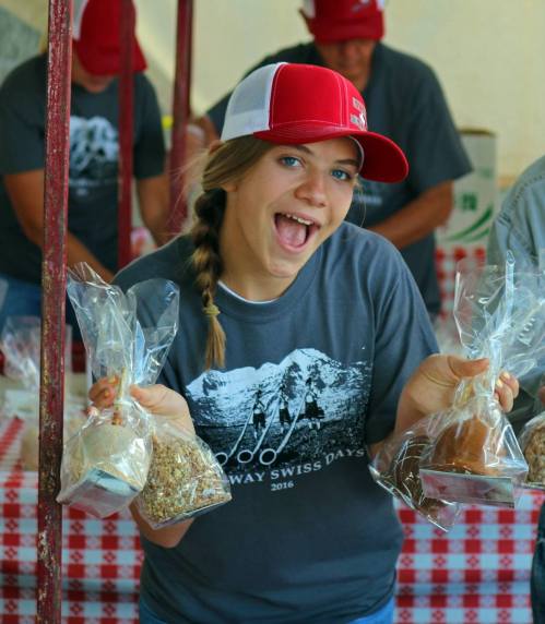 A smiling girl in a gray shirt and red cap holds bags of food at a festive outdoor event.