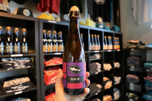 A hand holds a bottle of beer labeled "Soledera MV2" in front of shelves filled with clothing and other beer bottles.