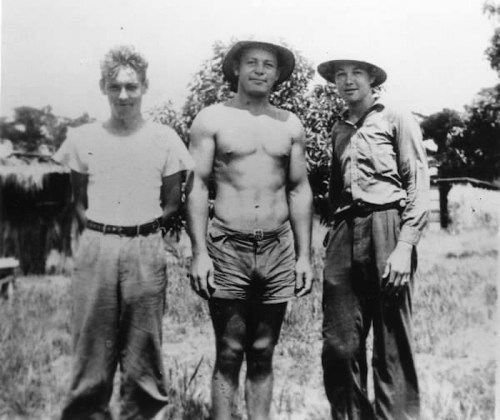 Three men stand outdoors in a grassy area, two wearing shirts and pants, one shirtless in shorts, all smiling.