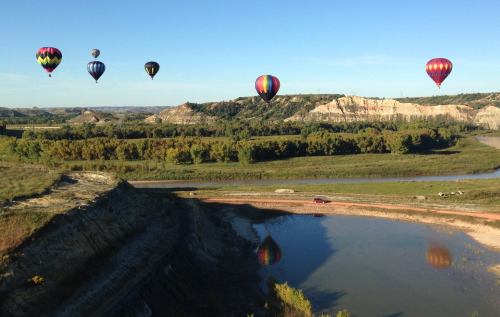 Colorful hot air balloons float over a scenic landscape with hills, trees, and a calm river reflecting the sky.