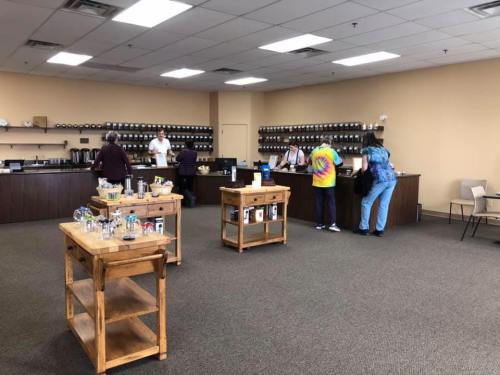 A spacious room with wooden tables, people interacting, and shelves filled with jars and containers in the background.