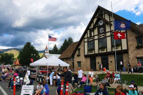 A crowd gathers outside a building with flags, enjoying an event on a sunny day with mountains in the background.