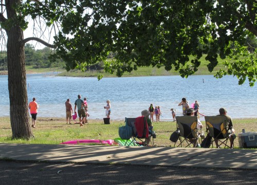 People enjoying a sunny day by the water, with chairs set up on the shore and trees providing shade.