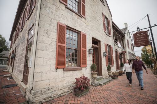 A couple walks past a historic stone building with red shutters and a sign, surrounded by potted plants and brick pavement.