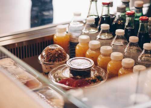 A display of various bottled drinks and snacks, including fruit cups and granola, in a glass case.