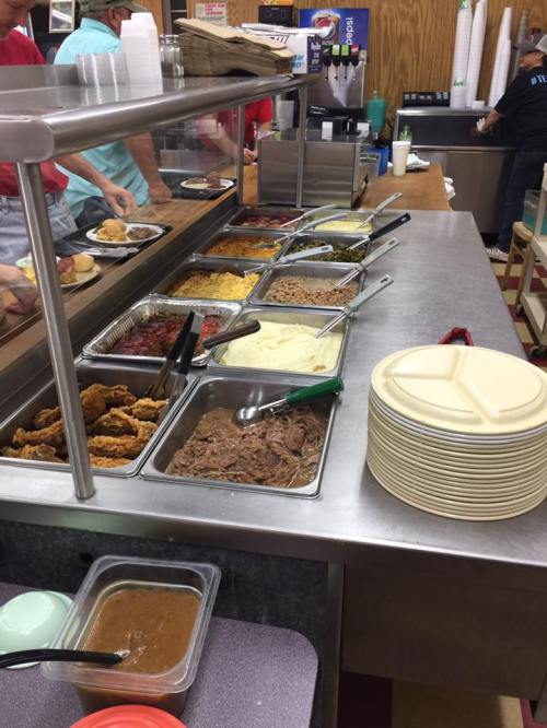 A buffet line with various dishes including meats, sides, and sauces, alongside stacks of plates.
