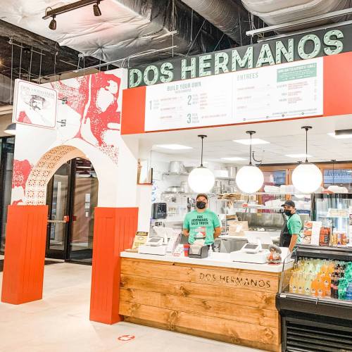 Interior of a restaurant named "Dos Hermanos," featuring a wooden counter, menu board, and staff in green shirts.