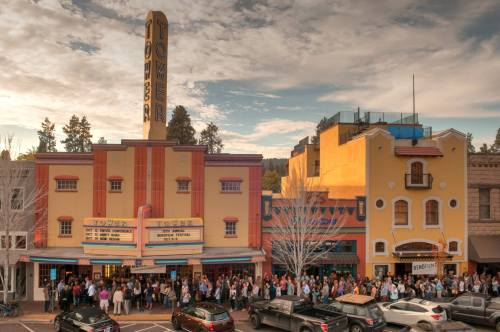 A crowd gathers outside the historic Tower Theater, with colorful buildings and a cloudy sky in the background.