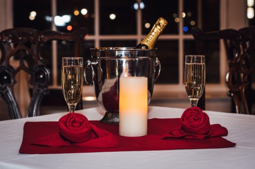 A romantic table setting with champagne, two glasses, a candle, and red rose decorations.