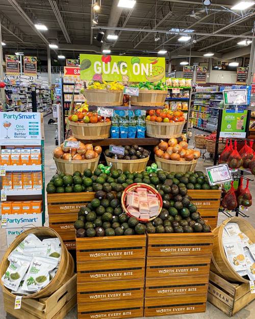 A colorful display of avocados, oranges, and drinks in a grocery store, with a sign that says "GUAC ON!"