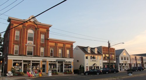 Historic brick buildings line a street, featuring shops and colorful chairs outside, under a soft evening sky.