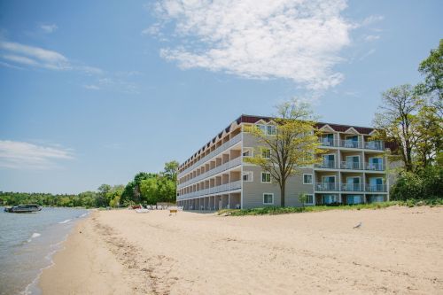 A beachside hotel with multiple balconies, surrounded by trees and a sandy shore under a clear blue sky.