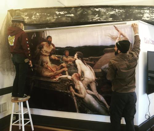 Two people hang a large painting depicting a dramatic scene on a wall, while a third person stands on a stool nearby.