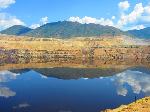 A scenic view of a mining site with layered earth, mountains in the background, and a reflective water surface.