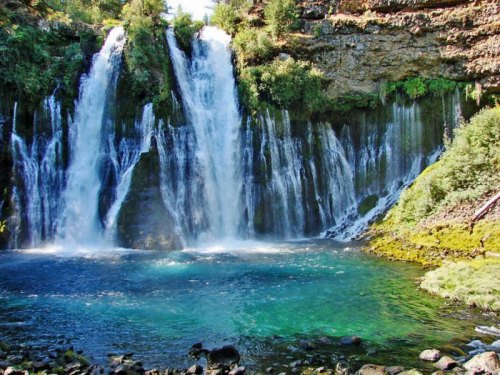 A scenic waterfall cascading into a clear blue pool, surrounded by lush greenery and rocky terrain.