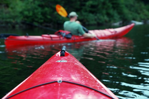 A close-up of a red kayak in calm water, with a person paddling in the background. Lush greenery surrounds the scene.