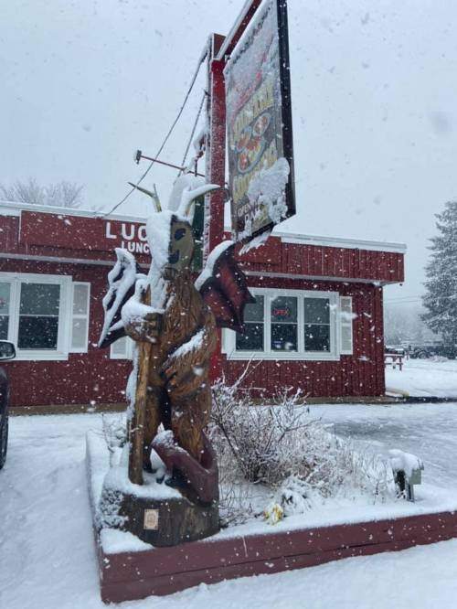 A wooden statue of a creature stands in front of a red building, covered in snow during a winter snowfall.