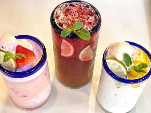Three colorful drinks in glasses: a pink strawberry drink, a dark red beverage with lime, and a creamy white drink with fruit.