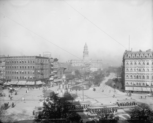 Historic city square with buildings, streetcars, and pedestrians, featuring a prominent clock tower in the background.