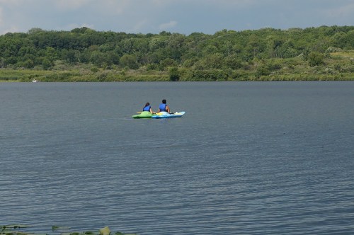 Two people kayaking on a calm lake surrounded by lush greenery under a partly cloudy sky.