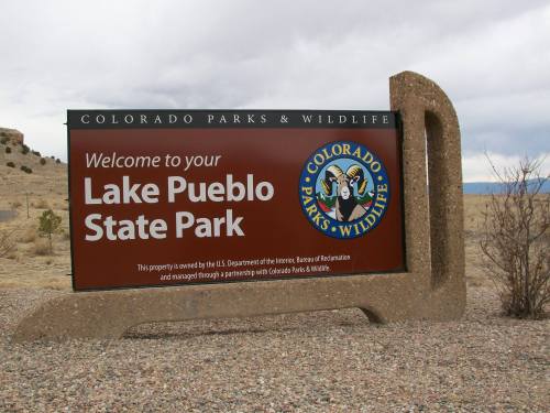 Sign welcoming visitors to Lake Pueblo State Park, featuring the Colorado Parks & Wildlife logo. Desert landscape in background.