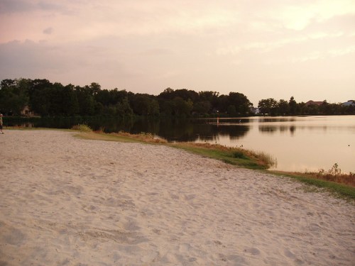 A serene lakeside view at sunset, featuring sandy shores and lush greenery reflecting in the calm water.