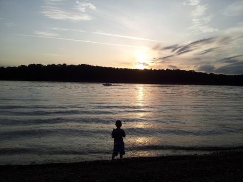 A child stands by the water's edge, watching a sunset over a calm lake with gentle waves.