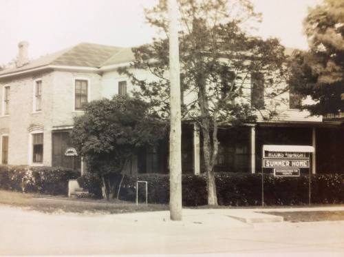 Black and white photo of a two-story house with a sign reading "SUMMER HOME" in front, surrounded by trees.