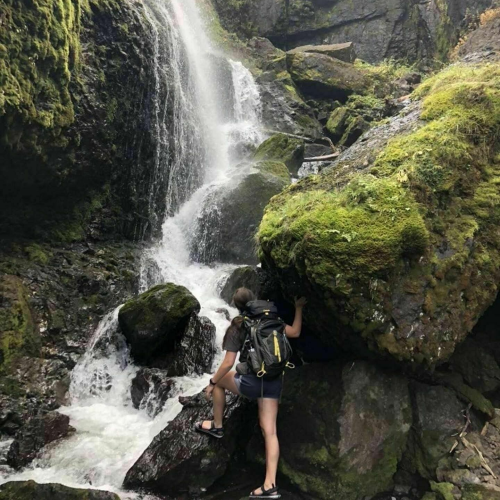 A person stands near a waterfall, surrounded by lush greenery and rocky terrain. Water cascades down the rocks.