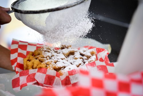 A hand sifts powdered sugar over a plate of funnel cake with a red and white checkered paper lining.