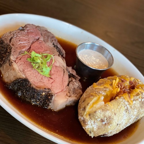 A plate featuring sliced roast beef with gravy, a baked potato topped with cheese, and a small cup of sauce.