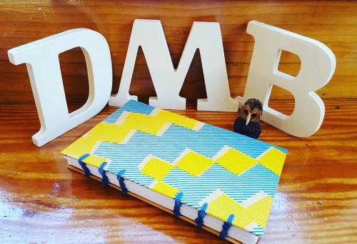 A colorful notebook with a geometric design, a small figurine, and white letters spelling "DMB" on a wooden surface.