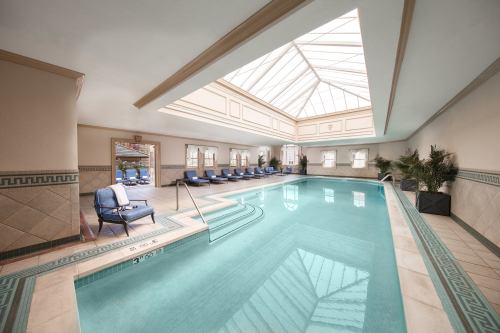Indoor swimming pool with a glass ceiling, surrounded by lounge chairs and potted plants. Bright and inviting atmosphere.