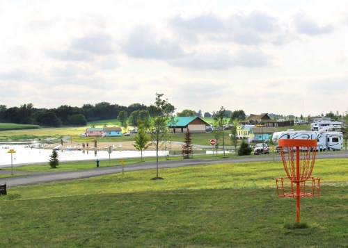 A scenic view of a park with a disc golf basket in the foreground, a beach area, and recreational vehicles in the background.
