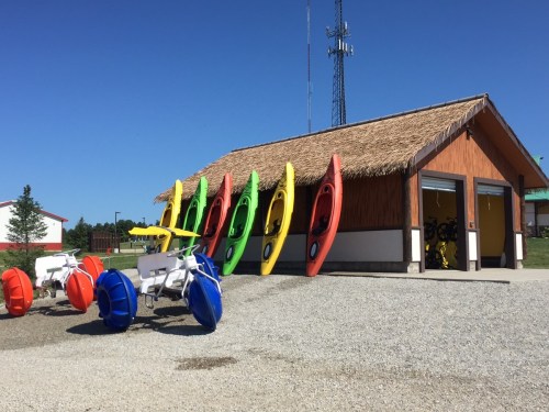 Colorful kayaks lined up outside a thatched-roof building, with pedal boats in the foreground on a sunny day.