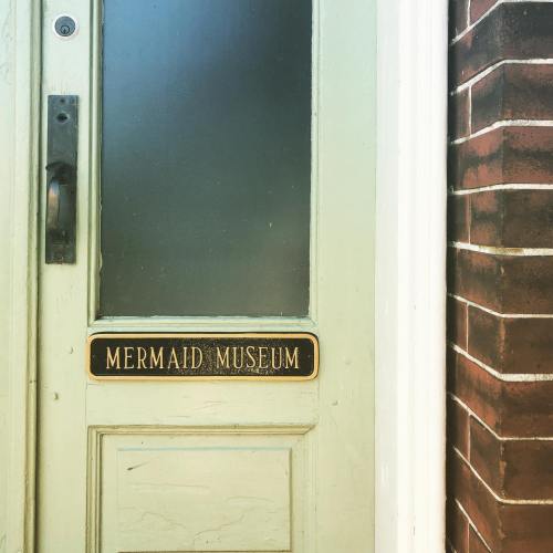 A door with a sign reading "Mermaid Museum," featuring a glass panel and a brick wall beside it.
