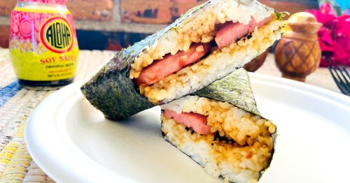 A close-up of a sushi sandwich with rice, seaweed, and layers of meat, next to a bottle of soy sauce.