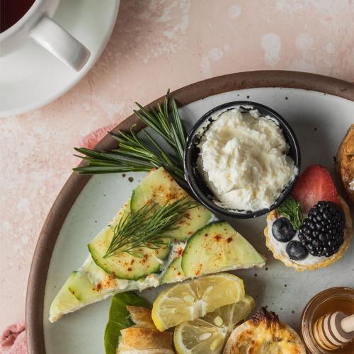 A plate with a slice of pie, fresh herbs, lemon, and a bowl of cream topped with berries, alongside a cup of coffee.
