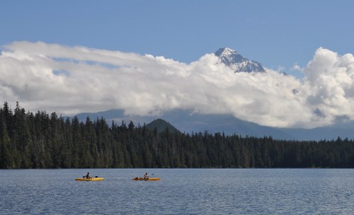 Two kayakers paddle on a calm lake surrounded by lush forests and a snow-capped mountain under a blue sky.