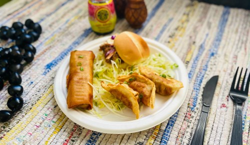 A plate with a spring roll, dumplings, a small bun, and shredded lettuce, set on a colorful woven tablecloth.