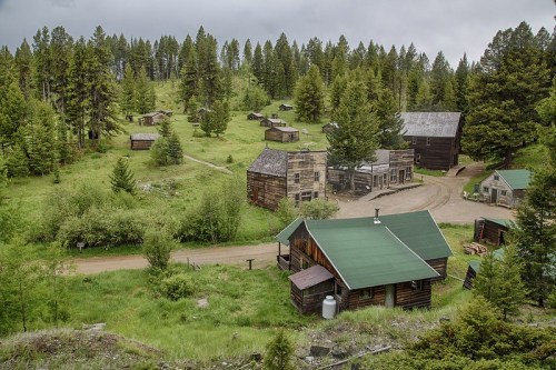 A scenic view of a rustic ghost town surrounded by lush greenery and tall trees under a cloudy sky.