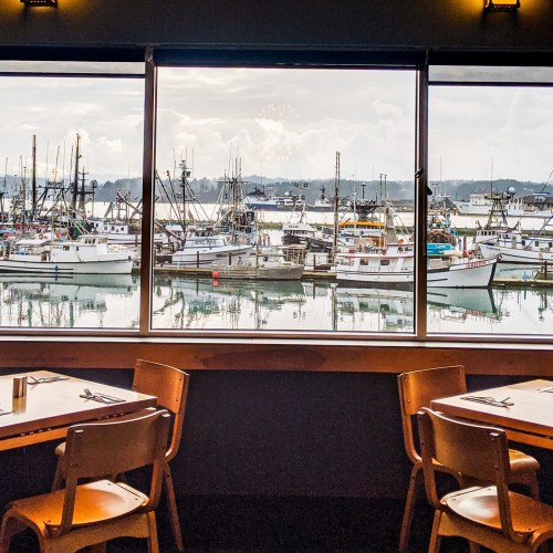 View from a restaurant window showing a harbor filled with fishing boats and a cloudy sky. Tables are set in the foreground.