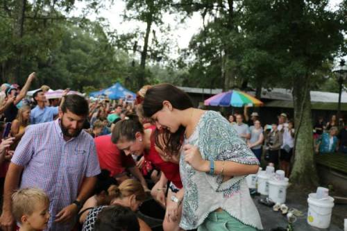 A lively outdoor event with a crowd gathered around participants engaging in a fun activity, surrounded by trees and tents.
